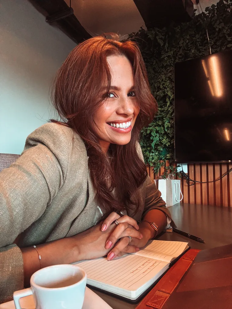 Rayane Guimarães sorrindo, sentada à mesa com um caderno e uma xícara de café